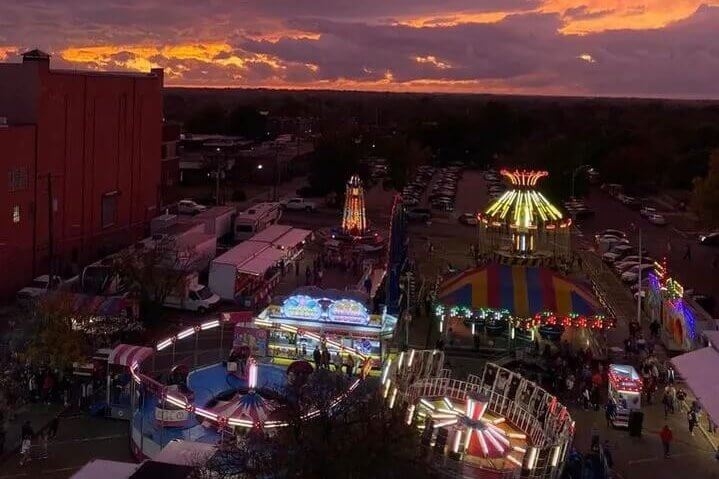 Aerial view of a carnival at dusk with colorful rides and lights beneath a dramatic pink and orange sunset sky.