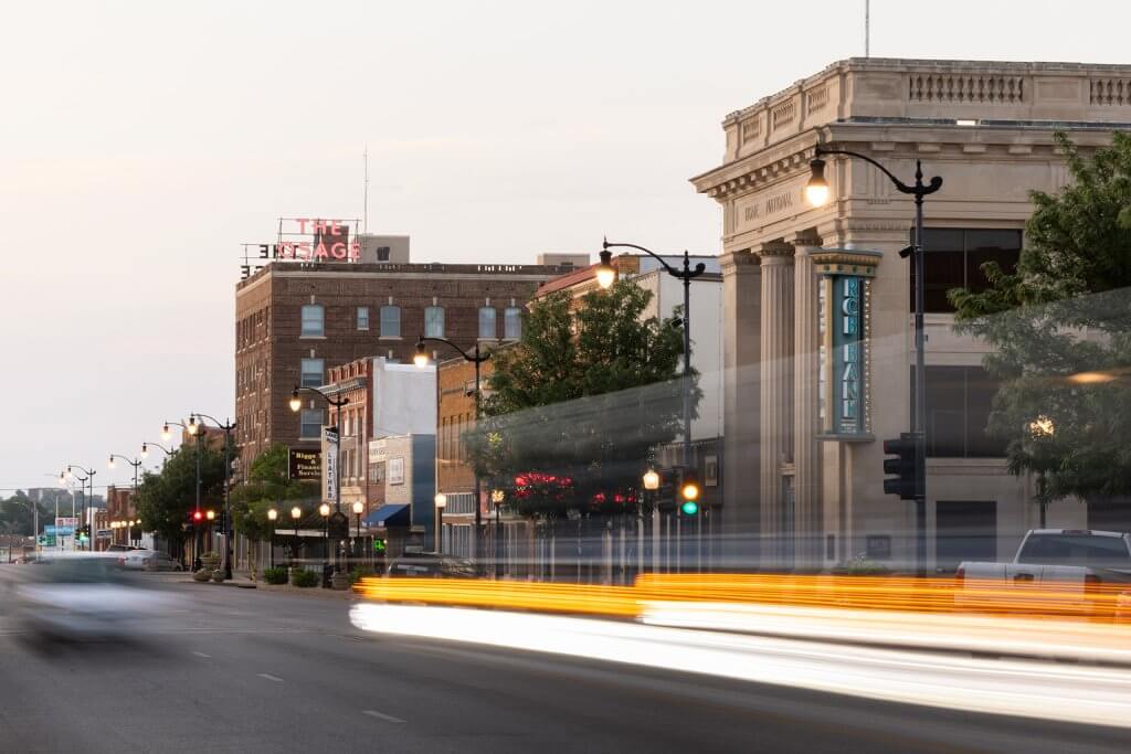 Downtown Arkansas City street at dusk with historic buildings, streetlights, and blurred car lights from passing traffic.