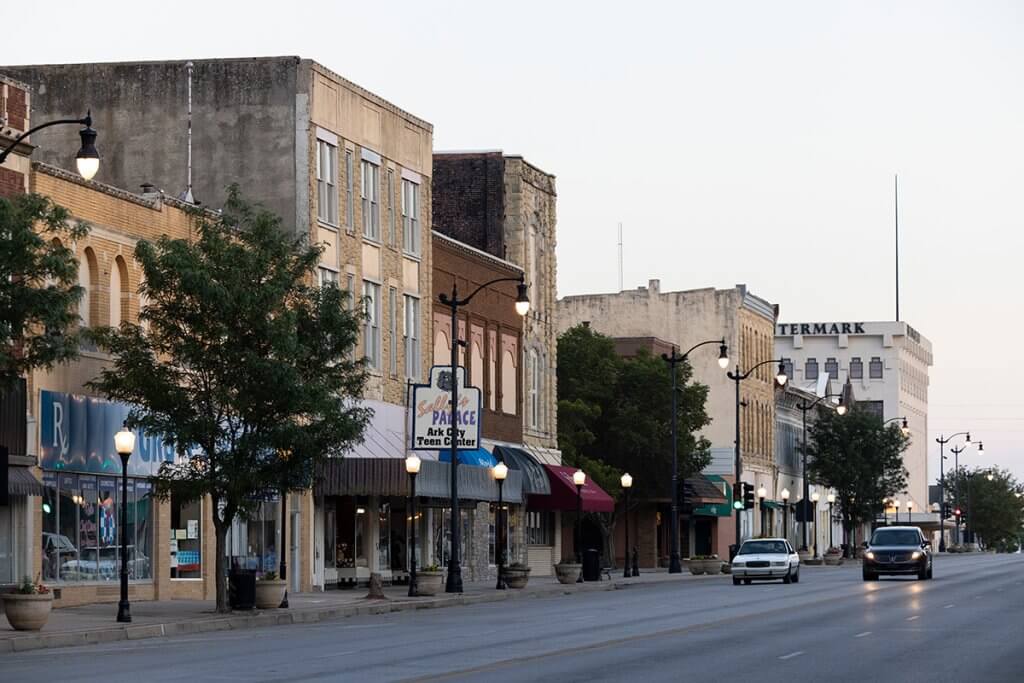 Historic downtown Arkansas City street with brick storefronts, streetlights, and light traffic at dusk.