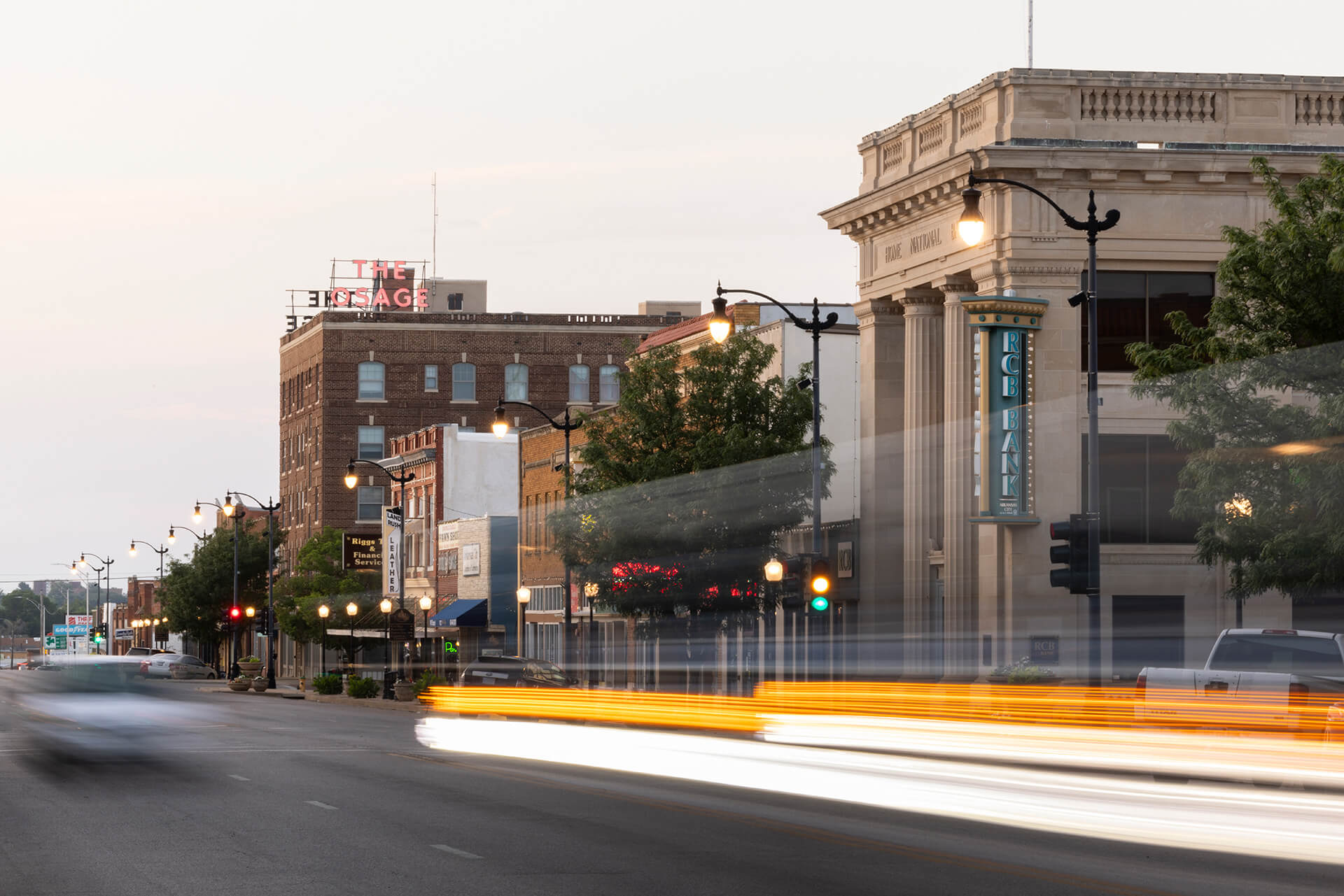 Downtown Arkansas City street at dusk with historic buildings, streetlights, and blurred car lights from passing traffic.