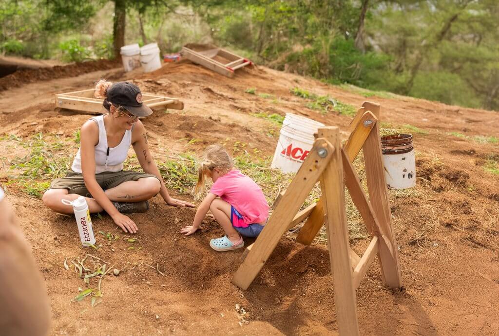An adult and a young child sit on the ground at an archaeology site, gently exploring soil together near sifting tools.