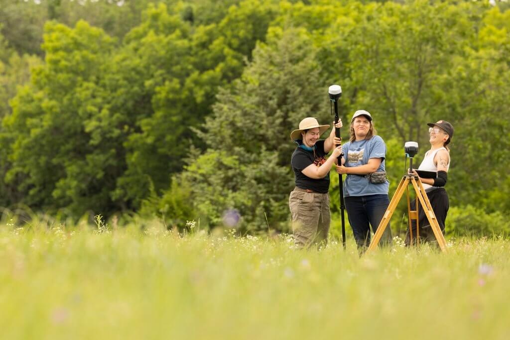 Three researchers stand in a grassy field using surveying equipment on tripods, with trees in the background.