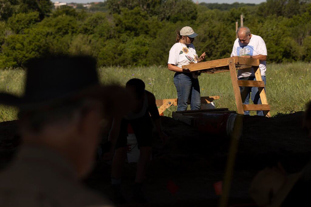 Two archaeologists examine soil on a wooden sifting screen in a grassy field during an excavation.