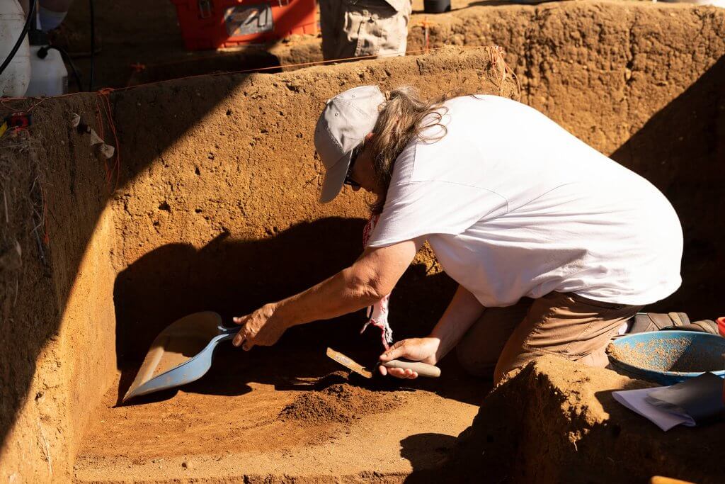 An archaeologist kneels in a trench, using hand tools to expose soil layers during excavation.
