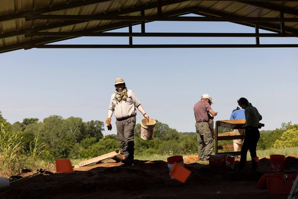 Archaeologists carry buckets and sift soil under a covered excavation shelter.