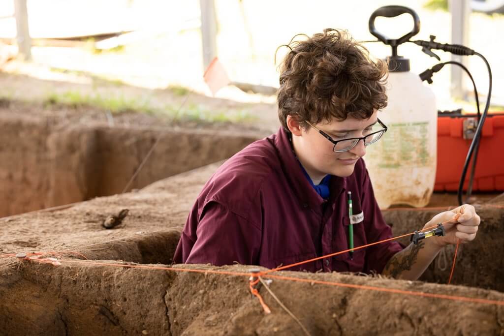An archaeologist measures and records a trench wall using string lines and a measuring tool.