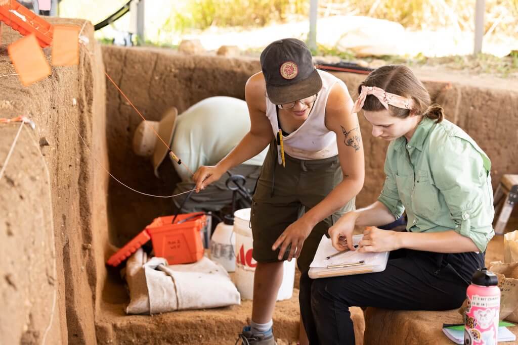Two archaeologists review notes and artifacts together beside an excavation trench.