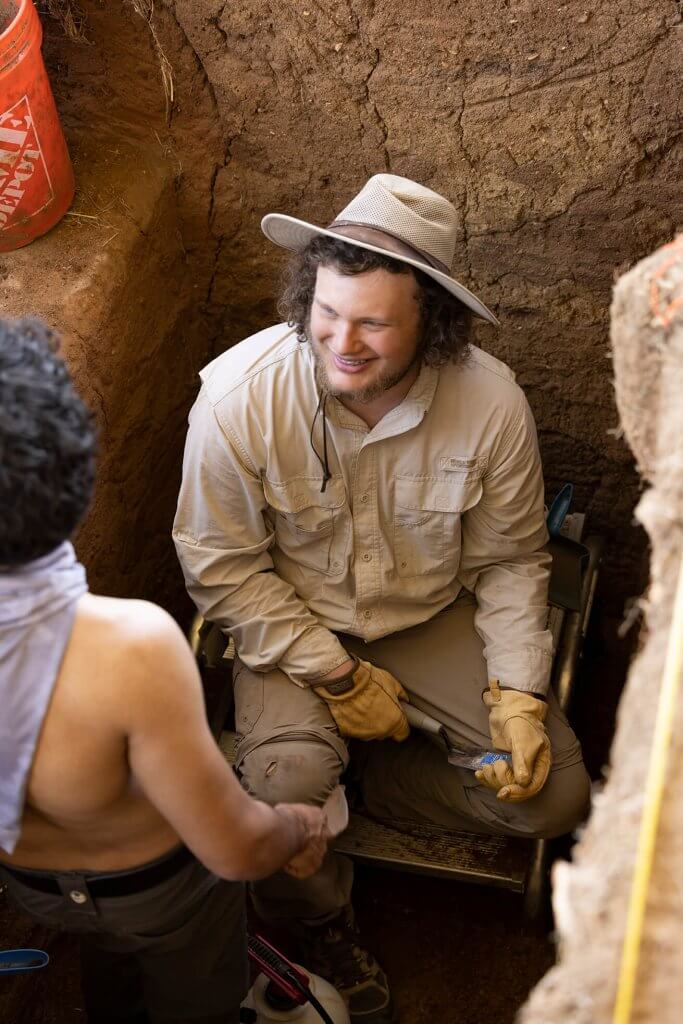 An archaeologist sits inside a trench smiling while speaking with another team member.
