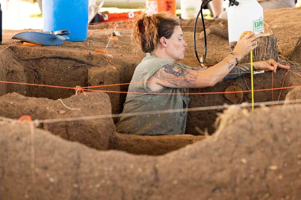 An archaeologist measures a trench depth using a tape measure while another records notes nearby