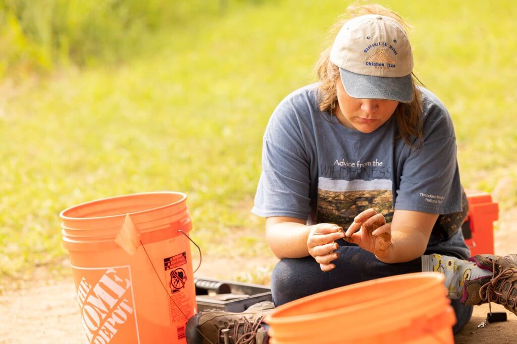 An archaeologist examines a small artifact while seated near buckets and excavation tools.