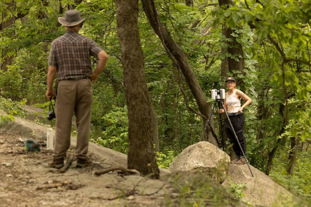 Two researchers use tripod-mounted scanning equipment in a wooded area during field documentation.
