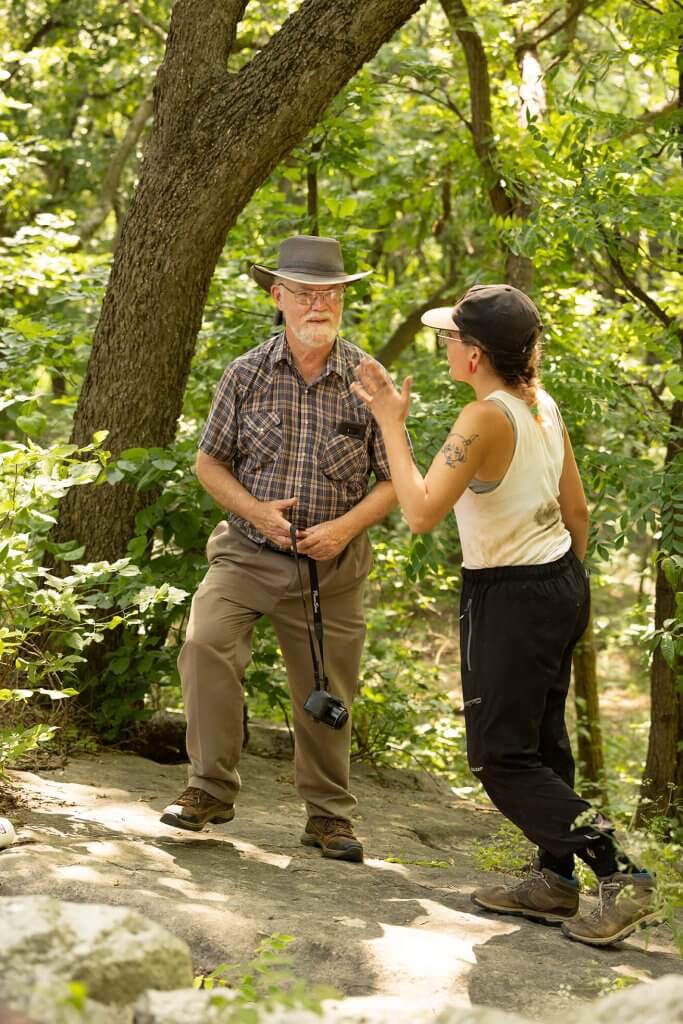 Two researchers talk on a wooded trail, one holding a camera while the other gestures during discussion.