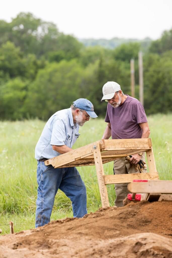 Two archaeologists examine soil on a wooden sifting screen in a grassy field.