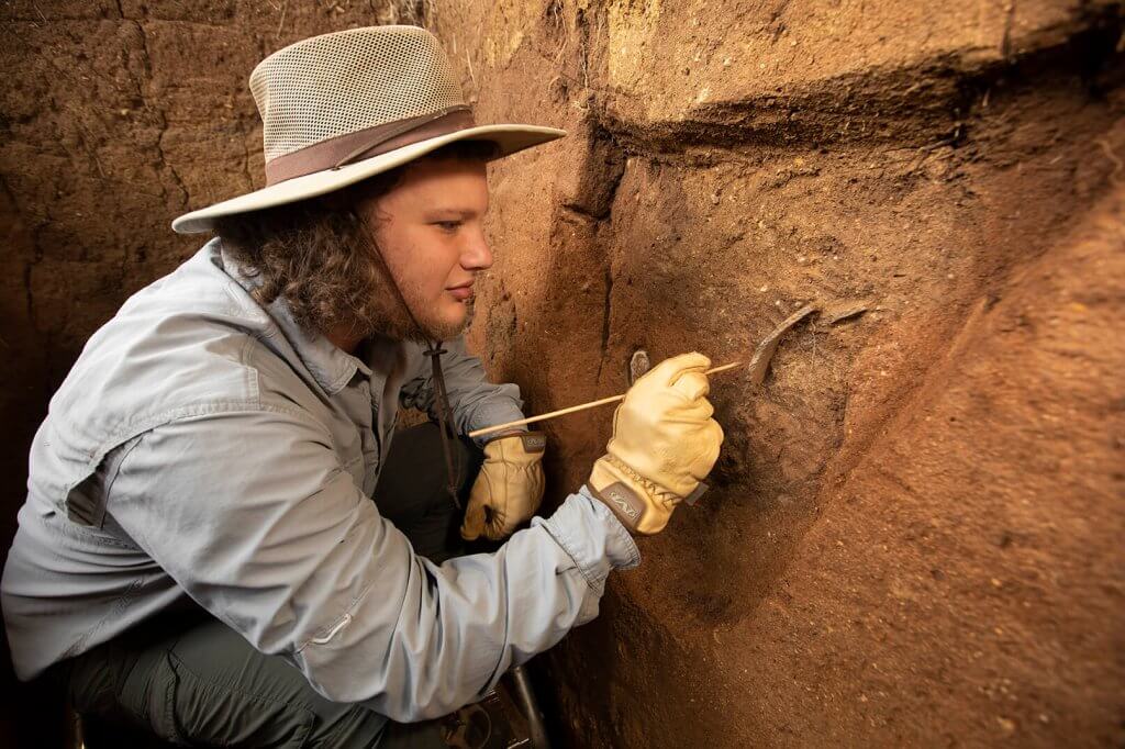 An archaeologist carefully cleans an exposed soil profile with a small tool inside an excavation unit.