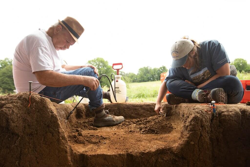 Two archaeologists kneel at an excavation trench, using hand tools to carefully remove soil.