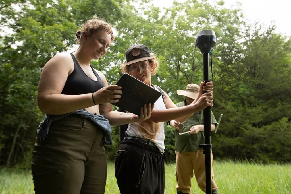 Two researchers review data on a tablet while standing beside surveying equipment in a grassy field.