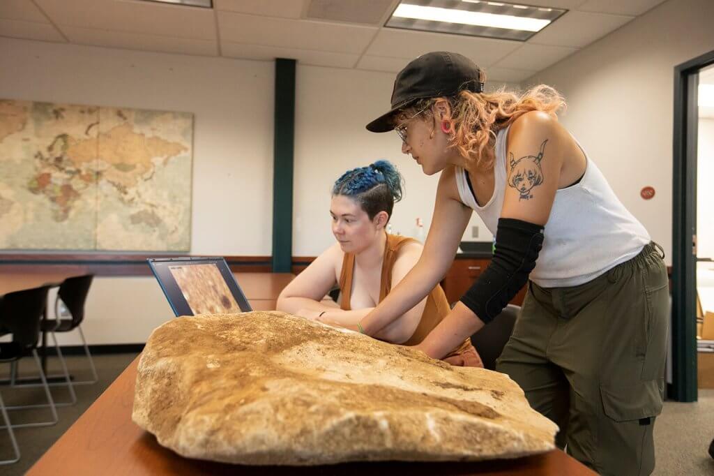 Two researchers examine a large stone artifact on a table inside a lab or classroom setting.