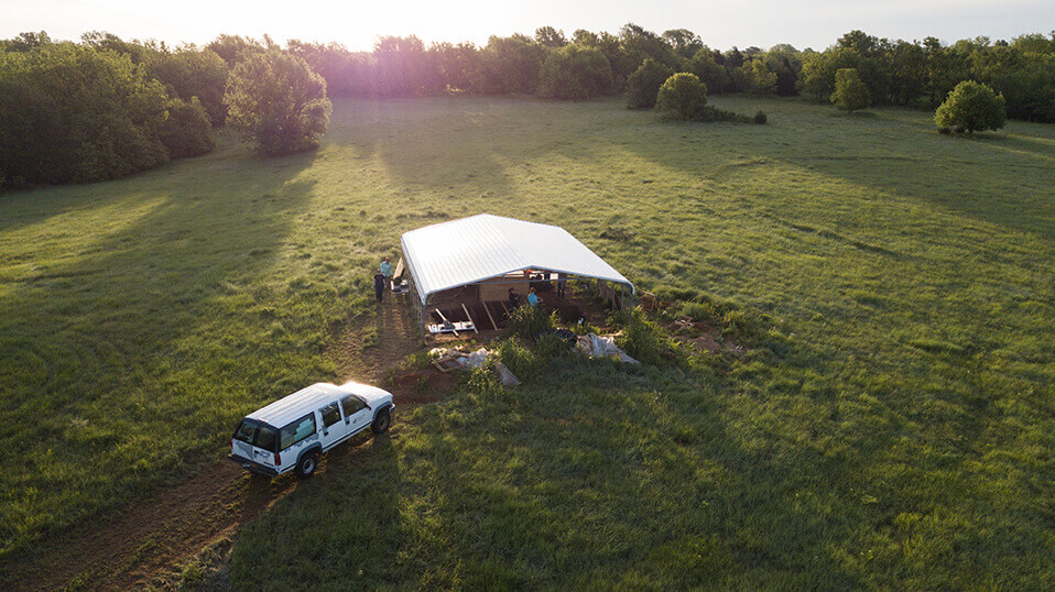 Aerial view of an excavation shelter and vehicle set in a wide grassy field at sunset.
