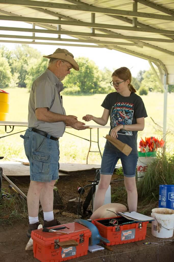 An archaeologist hands an artifact to a younger researcher beside labeled toolboxes at an excavation site.