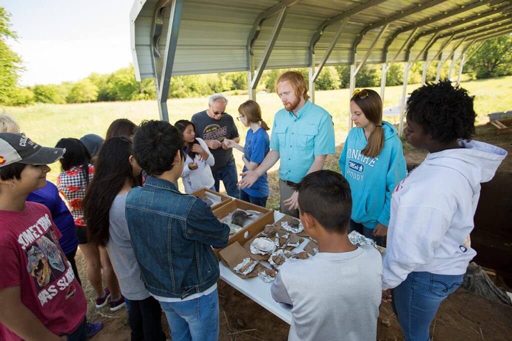 A group of students gathers under an open shelter while an instructor explains artifacts displayed on a table.