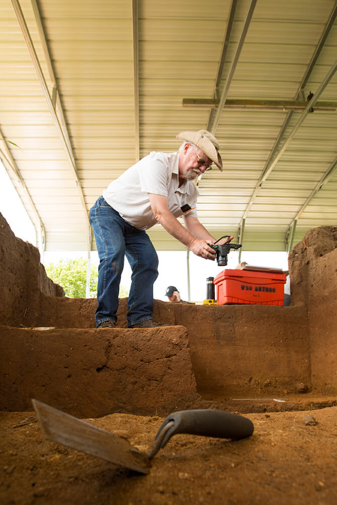 An archaeologist photographs soil layers inside an excavation trench under a covered shelter.
