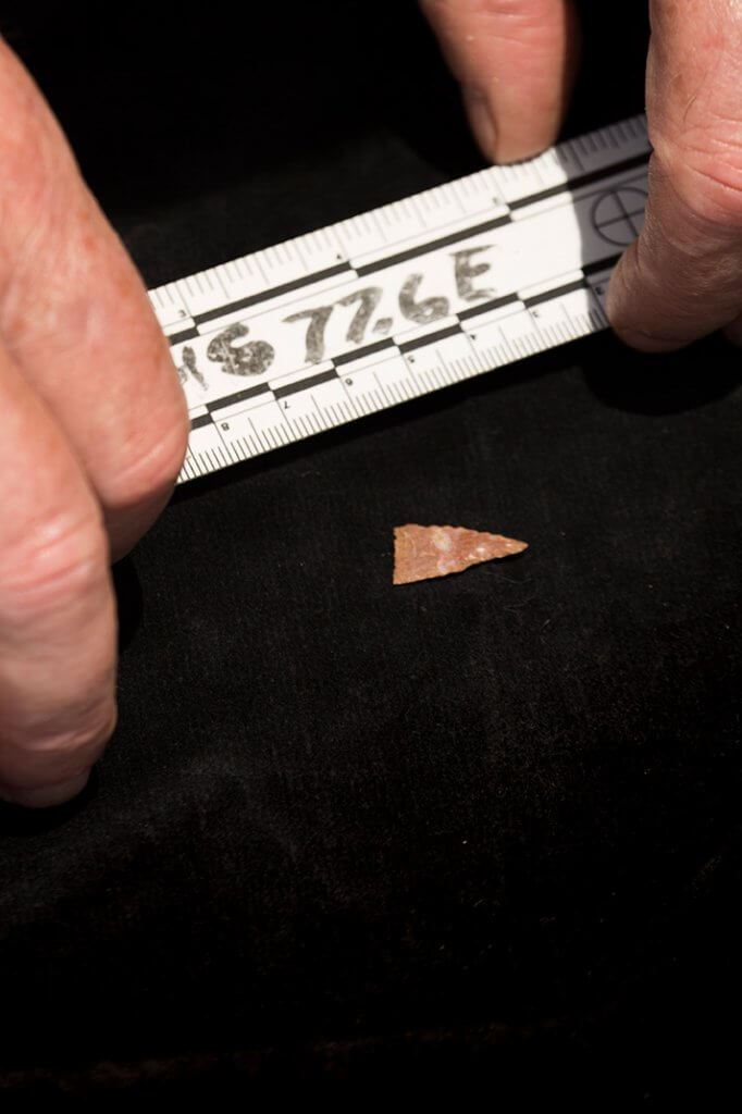 A small stone projectile point measured with a folding ruler against a dark background.