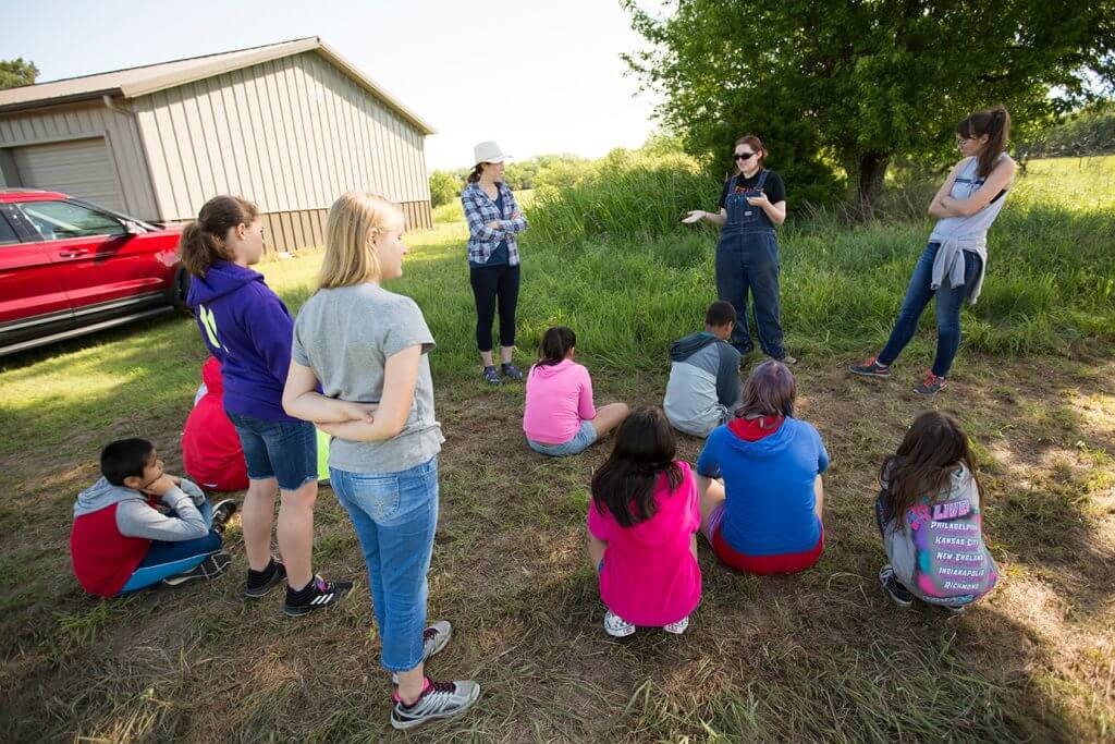 A group of children listens to an instructor during an outdoor archaeology lesson near a field building.