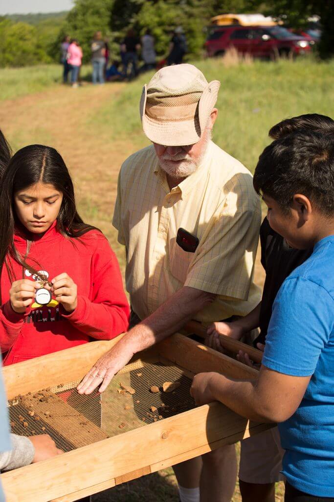 An instructor guides children as they examine soil and artifacts using a wooden sifting screen outdoors.