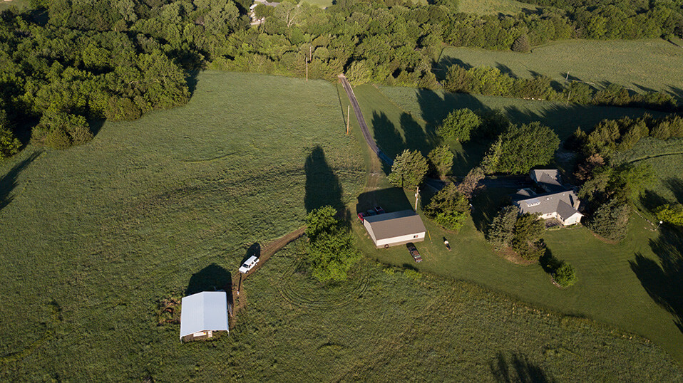 Aerial view of a rural landscape with grassy fields, trees, and several small buildings connected by dirt roads