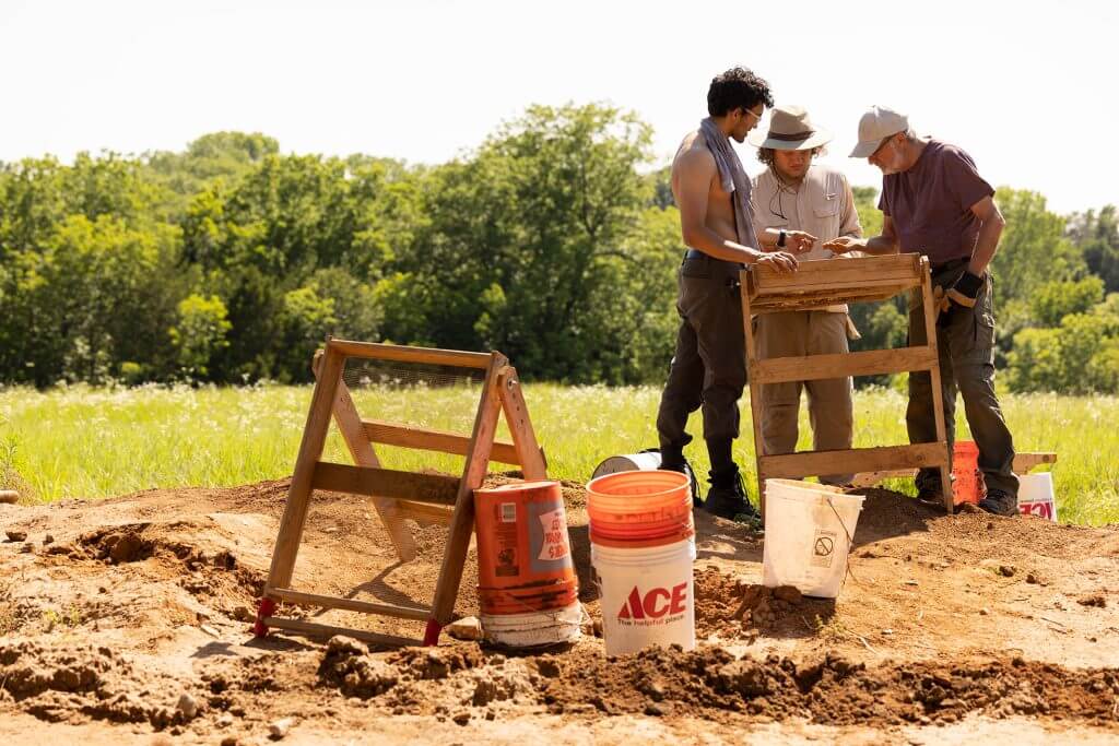 Archaeologists sift soil through wooden screens at an excavation site with buckets and tools nearby.