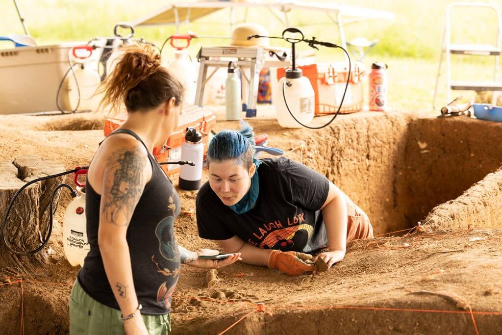 Two archaeologists examine soil layers and artifacts inside an excavation trench with equipment in the background.