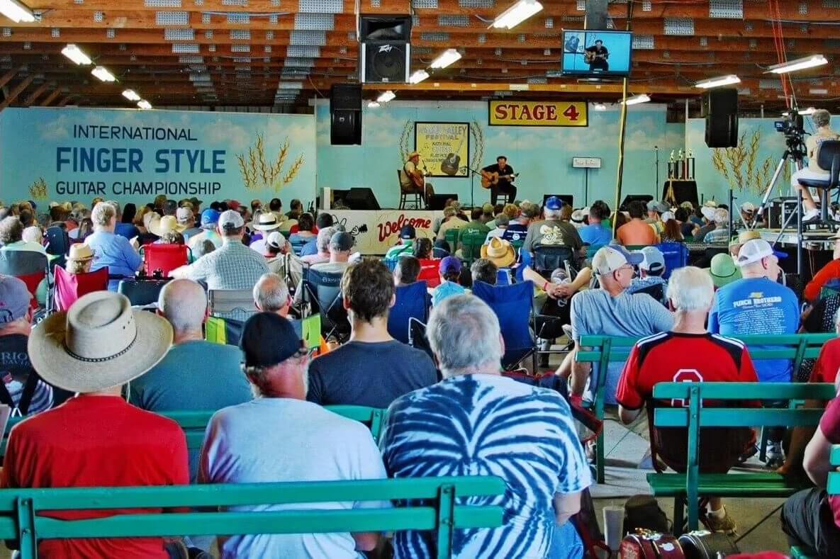 Audience seated on benches watches musicians perform on stage at the International Fingerstyle Guitar Championship inside a large pavilion.